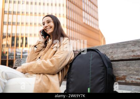 Brunette femme avec des lunettes assise sur un banc et parlant au téléphone Banque D'Images