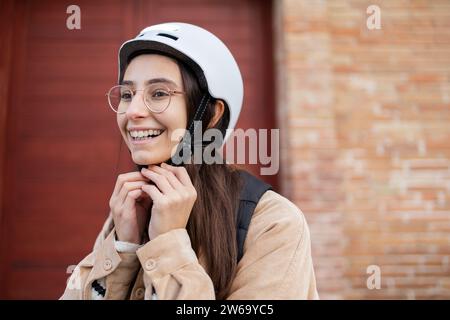 Une jeune femme joyeuse sécurise la sangle de son casque de vélo blanc devant un mur de briques, se préparant à une balade en toute sécurité. Banque D'Images