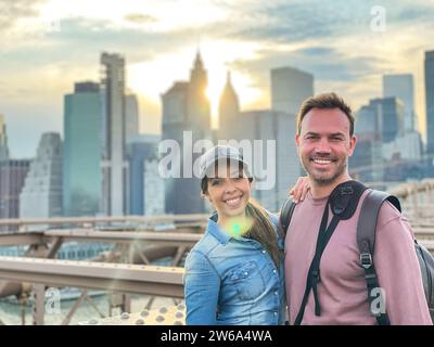 Un homme et une femme souriants debout ensemble sur le pont de Brooklyn avec l'horizon de Manhattan illuminé par le soleil couchant en arrière-plan. Banque D'Images