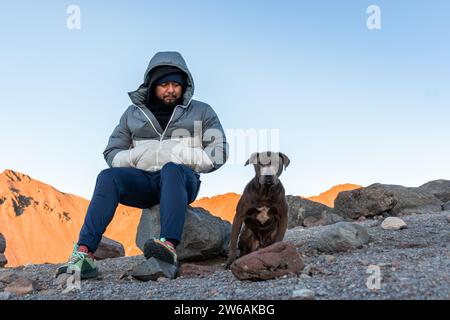 Barbu jeune randonneur mâle dans des vêtements chauds et la capuche regardant vers le bas tout en étant assis sur la roche sur la colline avec les mains dans les poches avec le chien animal de compagnie contre brillant Banque D'Images