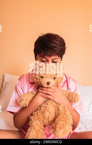 Jeune femme avec les cheveux courts et avec les yeux fermés tout en étant assise sur le lit en vêtements de nuit et embrassant l'ours en peluche doux dans les mains serrant contre orange Banque D'Images