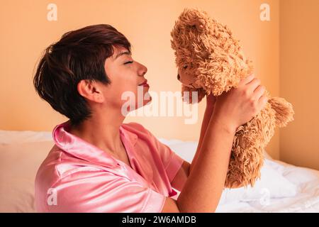 Vue latérale de la jeune femme avec les yeux fermés assis sur un lit confortable et tenant un petit ours en peluche dans la chambre Banque D'Images