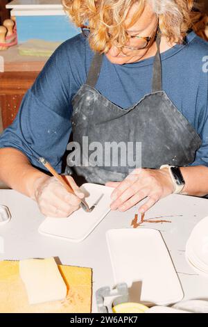 Concentré artisan féminin d'âge moyen dans des lunettes et tablier regardant vers le bas tout en étant assis à table avec divers articles en argile et en utilisant le pinceau à la déco Banque D'Images