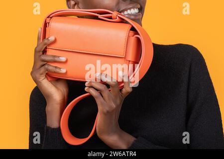 Heureux recadré anonyme jeune femme afro-américaine avec les cheveux courts tenant le nouveau sac à main orange tendance tout en regardant la caméra contre jaune b Banque D'Images