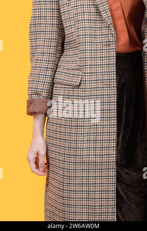 Femme mince caucasienne recadrée méconnaissable portant un long manteau à motif à carreaux regardant l'appareil photo tout en se tenant sur fond jaune Banque D'Images