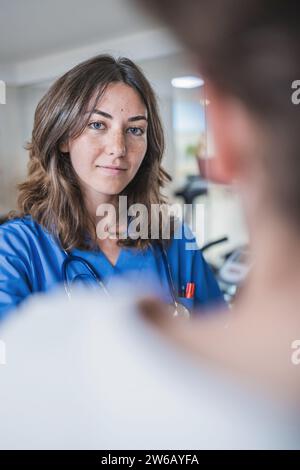 Souriant jeune femme médecin en uniforme bleu regardant la caméra tout en se tenant debout sur un fond flou dans le couloir de l'hôpital Banque D'Images