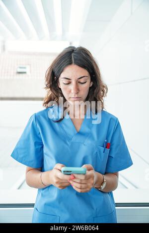 Jeune femme médecin portant l'uniforme bleu debout dans l'hôpital moderne et la navigation téléphone mobile Banque D'Images