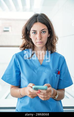 Portrait d'une jeune femme médecin portant un uniforme bleu debout dans un hôpital moderne et naviguant sur un téléphone mobile en regardant la caméra Banque D'Images