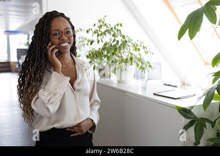 Pensive jeune femme d'affaires afro-américaine utilisant smartphone tout en se tenant dans le bureau souriant regardant atc amera Banque D'Images