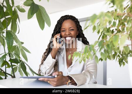 Jeune femme d'affaires afro-américaine joyeuse utilisant smartphone tout en naviguant sur tablette numérique au bureau dans le bureau regardant la caméra Banque D'Images