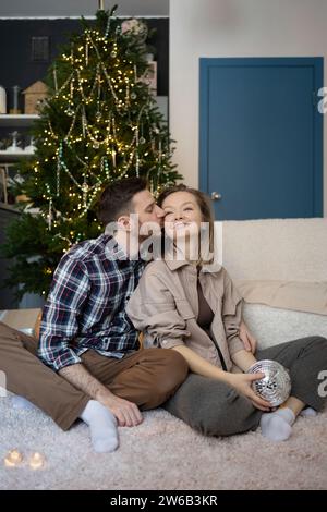 Jeune couple partage un moment d'affection avec un baiser affectueux sur le front. Ils sont assis confortablement dans leur maison, illuminés par la douce lueur d'un Banque D'Images