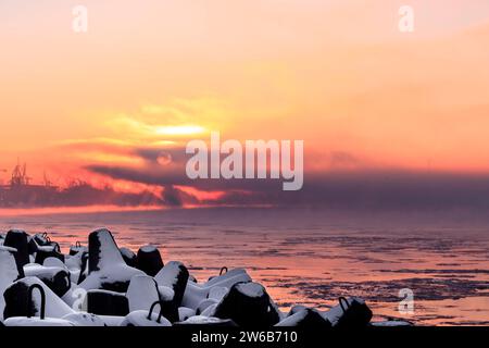 Brouillard matinal au-dessus de la neige recouvert de tétrapodes de béton le long de la côte avec des grues au loin, Klaipeda, Lituanie Banque D'Images