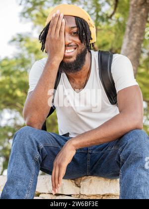 Souriant jeune homme afro-américain hipster portant un chapeau jaune et des écouteurs assis sur la frontière écoutant de la musique tout en couvrant la moitié du visage avec ha Banque D'Images