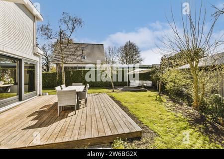 Maison moderne avec table à manger et chaises disposées sur la terrasse en bois par pelouse herbeuse contre le ciel bleu pendant la journée ensoleillée Banque D'Images