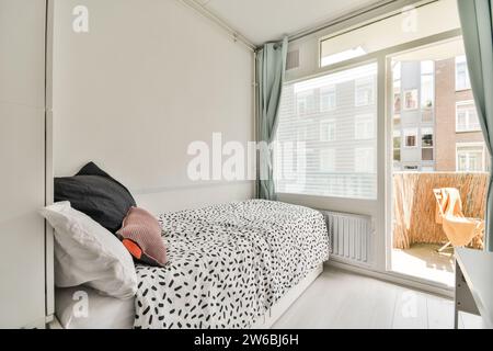 Chambre confortable et moderne avec lit simple et literie à motifs, grandes fenêtres offrant un éclairage naturel et vue sur le balcon. Banque D'Images