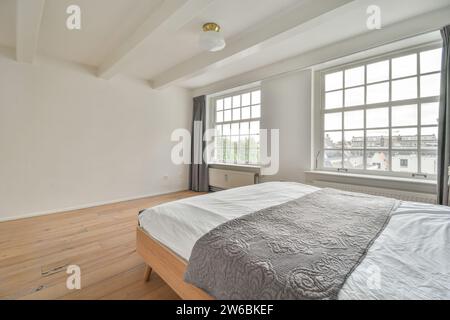 Cette chambre lumineuse et aérée dispose d''un grand lit avec couvre-lit gris, de parquet et de larges fenêtres permettant une lumière naturelle abondante. Banque D'Images