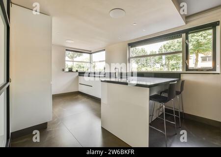 Une cuisine moderne élégante dispose d'un bar à petit-déjeuner, de grandes fenêtres et des meubles blancs sur un comptoir sombre. Banque D'Images