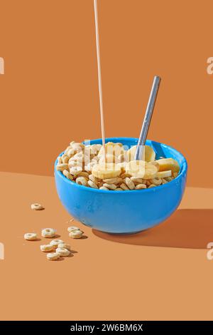 Un jet de lait étant versé dans un bol de céréales bleu rempli de cornflakes et de tranches de banane, sur fond orange. Banque D'Images