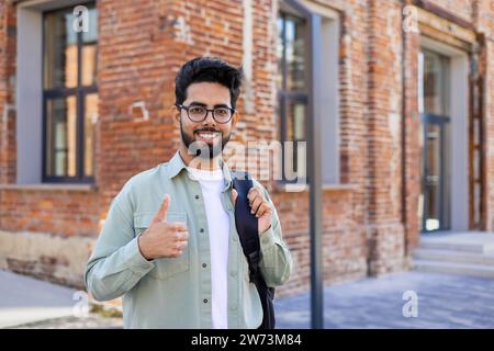 Portrait d'un jeune étudiant indien, freelance debout dehors dans la rue avec un sac à dos, souriant à la caméra et montrant le super signe avec son doigt. Banque D'Images
