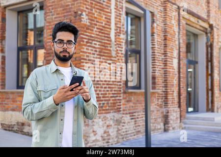 Portrait d'un Indien sérieux et confiant debout dans la rue à l'extérieur d'un bâtiment, tenant un téléphone portable et regardant l'appareil photo. Banque D'Images