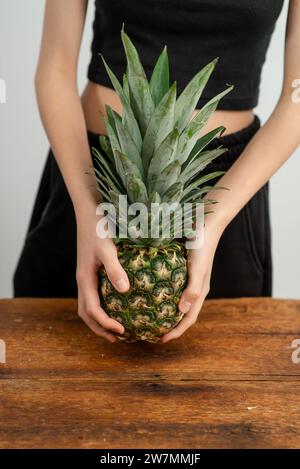 Gros plan d'une personne méconnaissable tenant un ananas mûr, posé sur un fond neutre sur une surface en bois rustique. Banque D'Images
