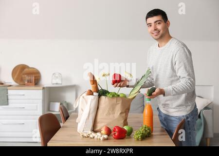 Jeune homme prenant des légumes frais de sacs d'épicerie à table dans la cuisine Banque D'Images