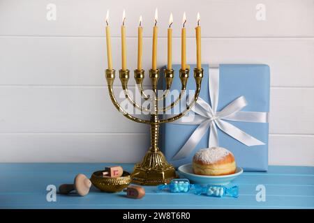 Célébration de Hanukkah. Menorah avec des bougies allumées, dreidels, beignet et boîte-cadeau sur la table en bois bleu clair Banque D'Images