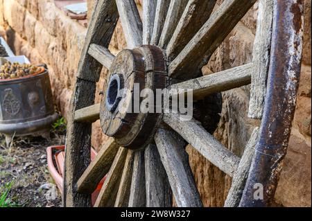 Vieille roue en bois avec des rayons d'un chariot mulet, vu de côté, appuyé contre le mur. La jante en fer recouvrant le bois sur la bande de roulement est visible. Banque D'Images