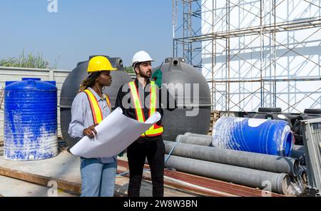 Deux ouvriers de la construction sont debout ensemble devant un grand chantier de construction avec casque et équipement de sécurité. Banque D'Images