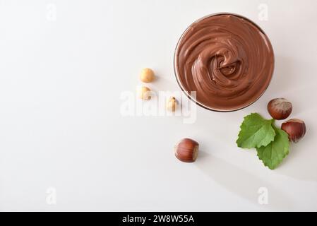 Crème de cacao dans un bol en verre avec noisettes et feuilles autour isolé sur table blanche. Vue de dessus. Banque D'Images