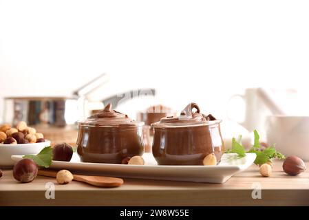 Deux bocaux en verre pleins de crème de cacao avec des noisettes sur une plaque avec des feuilles et des noix autour sur un banc de cuisine en bois blanc fond isolé. Vue avant. Banque D'Images