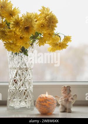 Gros plan vertical de chrysanthèmes jaunes dans un vase en verre, bougie allumée et statue en céramique d'un ange sur un rebord de fenêtre blanc avec panorama forestier. Banque D'Images