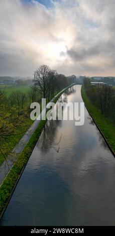 Cette image verticale présente une scène tranquille d'un canal s'étendant au loin, flanqué de rangées d'arbres et d'un chemin d'un côté. La lumière tôt le matin filtre à travers un ciel nuageux, projetant une douce lueur sur l'eau et créant une surface réfléchissante qui reflète le ciel et les arbres. L'herbe verdoyante sur les berges et les feuilles bourgeonnantes sur les arbres signalent le renouveau du printemps. Ce cadre paisible transmet un sentiment d'immobilité et la beauté d'une nouvelle journée. Canal serein au lever du soleil. Photo de haute qualité Banque D'Images