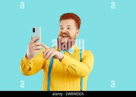 Heureux homme barbu gras dans la tenue à la mode enregistrement vidéo ou prise de selfie sur téléphone portable Banque D'Images