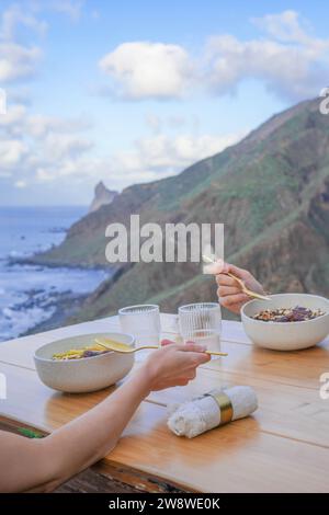 Une scène intime de dîner en plein air, mains servant de la nourriture, avec un océan à couper le souffle et la montagne en toile de fond à la lumière du jour Banque D'Images