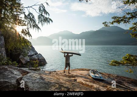 L'homme porte la planche à pagaie sur la tête après une pagaie tôt le matin. Banque D'Images