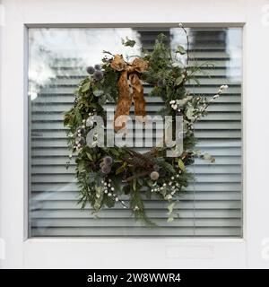 Couronne de Noël en branches de sapin, genévrier, lierre, saule, hellébore, eryngium, avec un arc en or sur une porte vitrée à Londres Banque D'Images