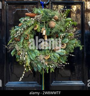 Couronne de Noël en branches de sapin, genévrier, saule, avec cônes de sapin, cercles oranges séchés, cloches et mousse sur une porte noire de Londres Banque D'Images