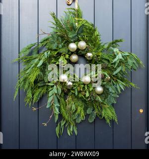 Une modeste couronne de Noël faite de brins de sapin, thuja, eucalyptus et boules d'argent sur une porte gris foncé à Londres. Cadre carré. Banque D'Images