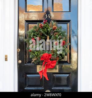 Couronne de Noël faite de branches de sapin, de pommes de pin, de baies de viburnum rouges et d'un arc sur une porte noire de Londres. Cadre carré. Banque D'Images