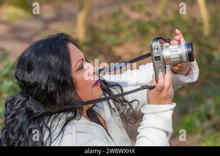 Femme d'âge moyen très concentrée prenant une photo avec son appareil photo numérique dans le parc public, fond brun flou avec des reflets verdâtres, da ensoleillé Banque D'Images