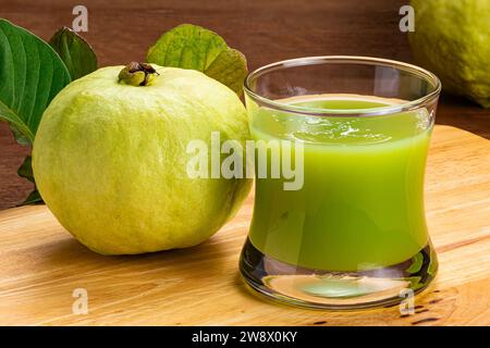 Vue en gros plan du jus de goyave vert frais en verre transparent et fruits de goyave doux mûrs avec des feuilles sur une planche de bois. Banque D'Images