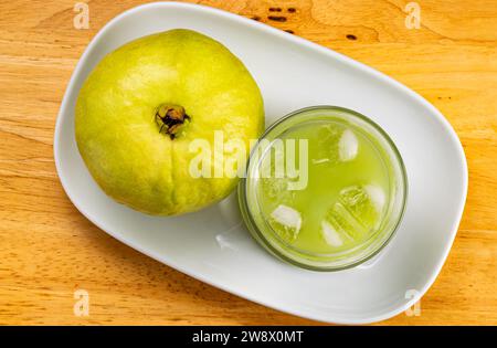 Vue de dessus du jus de goyave doux froid frais en verre transparent et fruits de goyave frais mûrs dans un plateau en céramique blanche sur une table en bois. Banque D'Images