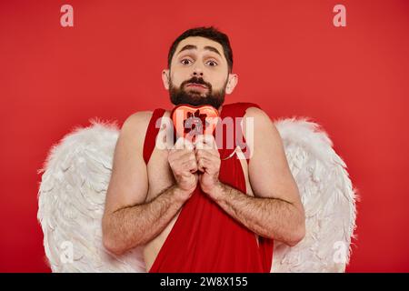 homme drôle en costume de cupidon et ailes tenant la boîte cadeau en forme de coeur et regardant la caméra sur rouge Banque D'Images