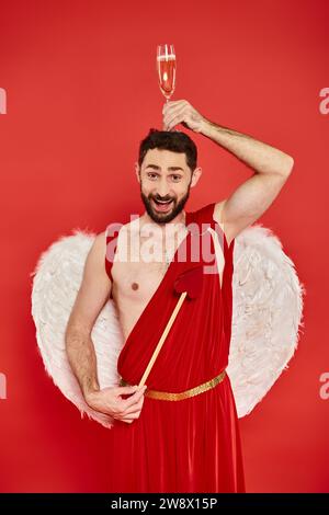 homme drôle et joyeux en costume de cupidon posant avec la flèche en forme de cœur et le verre de champagne sur la tête Banque D'Images