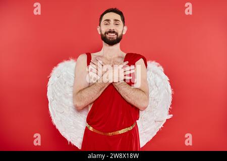 heureux homme barbu en costume de cupidon debout avec les mains sur la poitrine et regardant la caméra sur le rouge Banque D'Images