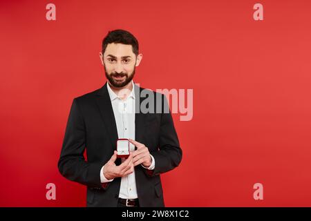 homme barbu souriant en costume noir montrant bague dans une boîte à bijoux sur rouge, st valentin présent Banque D'Images