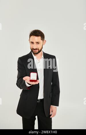 homme élégant joyeux montrant boîte à bijoux avec bague regardant la caméra sur gris, st valentines concept Banque D'Images