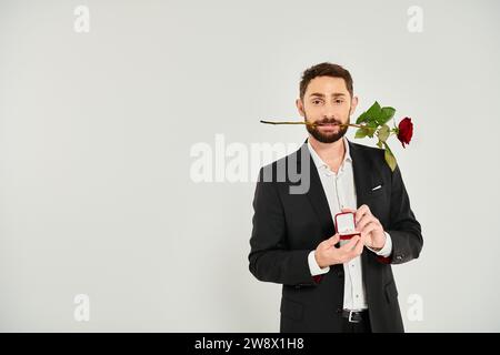 Bel homme avec rose rouge dans les dents montrant boîte à bijoux sur gris, Saint Valentin présent Banque D'Images