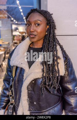 Une femme africaine élégante se tient en toute confiance dans un cadre urbain pendant la soirée. Elle est habillée à la mode dans une veste en cuir noir avec un col en rason blanc, complétant ses longs dreadlocks tordus. La toile de fond présente un café avec un éclairage chaleureux qui illumine doucement son visage, et des jeux de lumières ajoutent un effet bokeh à la scène. Le maquillage de la femme est fait avec goût, mettant en évidence sa beauté naturelle et son comportement posed. Son regard est dirigé vers la caméra avec un léger sourire assuré, suggérant à la fois l'accessibilité et l'assurance de soi. Urban chic Black Girl. Haute qualité Banque D'Images